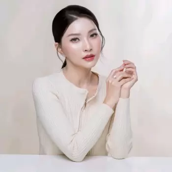wanita, cantik, muda, asia, potret, model, wajah, rambut hitam, sanggul, riasan, bibir merah muda, mata hijau, kalung, sweater, krem, lengan panjang, kerah bulat, ritsleting, cincin, tangan, jari, pose, duduk, meja putih, latar belakang polos, studio, profesional, elegan, lembut